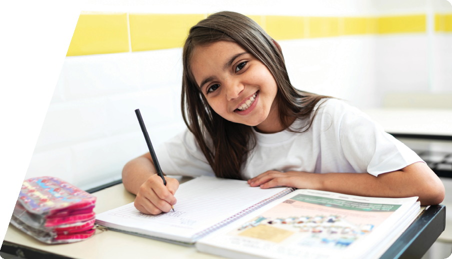 Foto de menina de cerca de dez anos, pele clara, cabelos lisos compridos e soltos. Ela olha para a câmera e sorri. Sentada em uma carteira escolar, segura um lápis sobre um caderno e um livro.